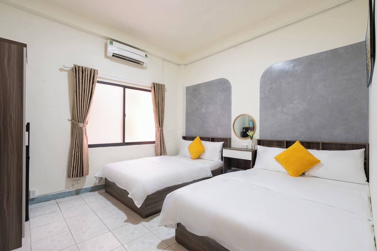 Hotelkamer met twee bedden, een raam met gordijnen, een airconditioning, een nachtkastje met spiegel en een klein bureau met stoel.