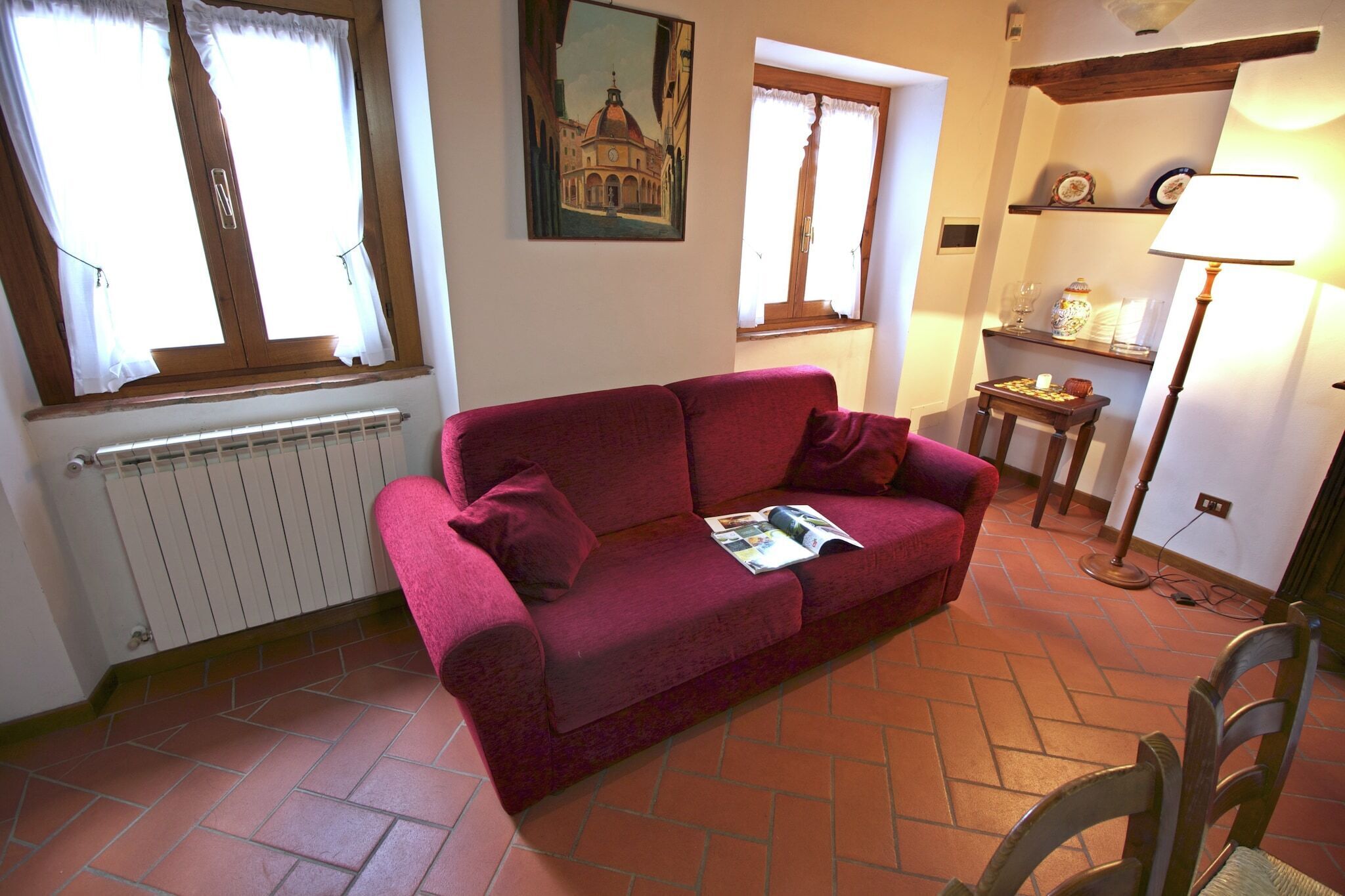 A living room with a red sofa, two windows with curtains, a radiator, a table with a lamp, and a chair.