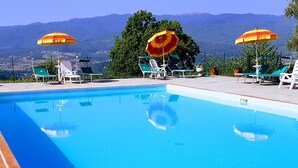 A swimming pool with lounge chairs and umbrellas, surrounded by greenery and mountains in the background. - Apartment in Tuscany near Vineyards (Poppi)