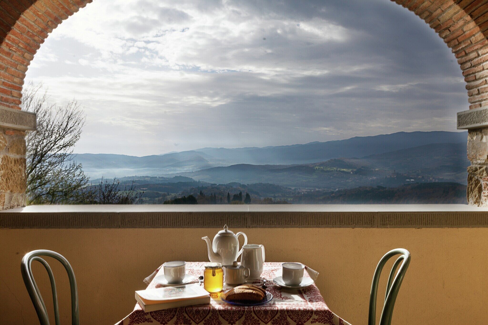 A table with a teapot, cups, and a book, overlooking a mountainous landscape through an arched window.