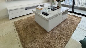A modern living room with a marble wall, a flat-screen TV, a white console table with a remote, a vase of flowers, and a large window with a city view.
