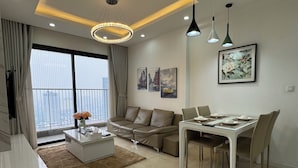 A modern living room with a sofa, dining table, and chairs. There is a large window with curtains, a chandelier, and artwork on the wall.