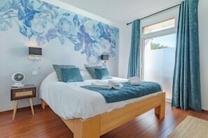 Ein Schlafzimmer mit einem Bett, einem Wandventilator, einem Nachttisch und einem Fenster mit VorhÀngen.