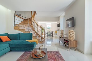 Ein modernes Wohnzimmer mit einem türkisblauen Sofa, einer Holztreppe, einem Flachbildfernseher und einem Essbereich im Hintergrund.