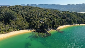 An aerial view of a coastal area with a beach, forested hills, and a small building on a cliff. - Jacuzzi Suite - Ocean Views & Beach (Kaiteriteri)