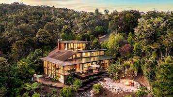 A modern two-story house with a glass facade, surrounded by dense forest.