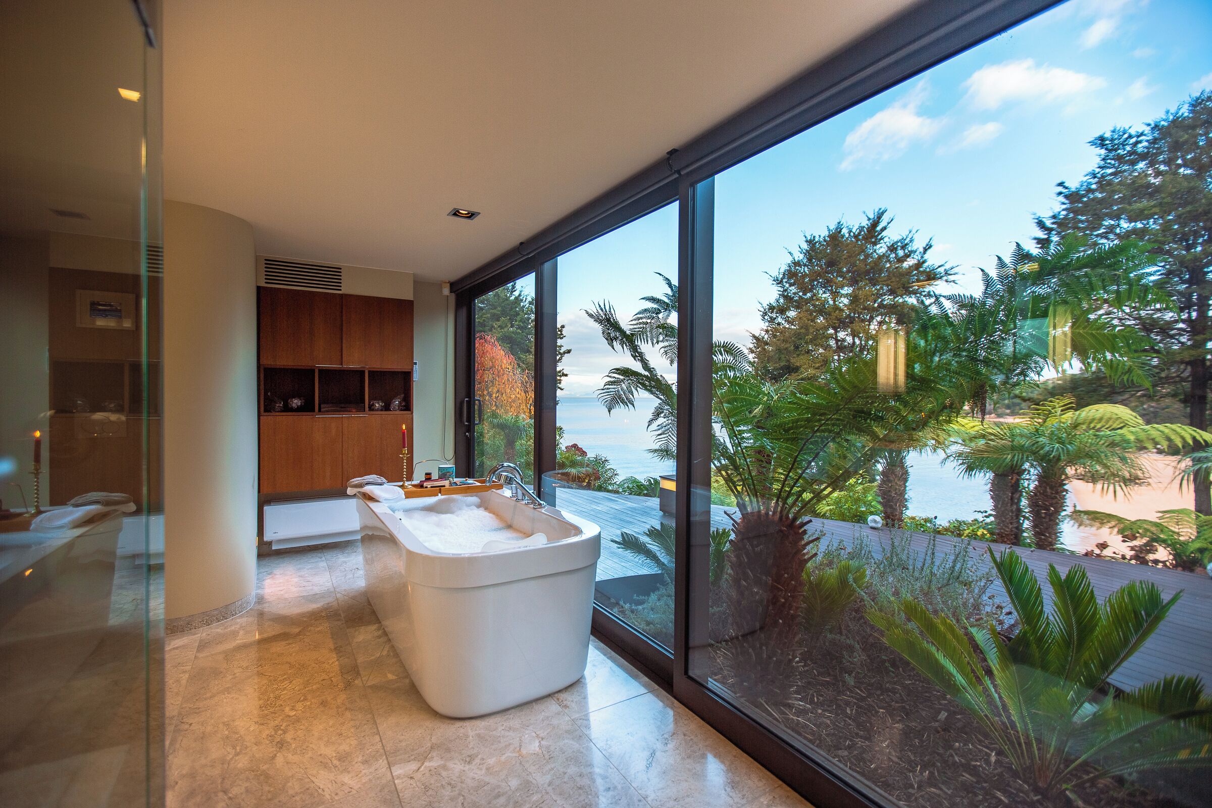 A modern bathroom with a large bathtub, a glass shower enclosure, and a view of a tropical garden and ocean.
