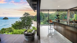 A modern interior with a large glass door leading to a balcony overlooking a beach and ocean. - Jacuzzi Suite - Ocean Views & Beach (Kaiteriteri)