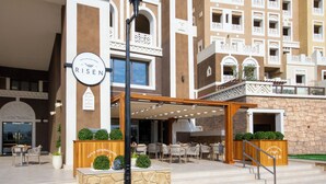 L’entrée d’un hôtel, avec une enseigne indiquant « RISE », un espace extérieur aménagé avec des chaises en osier et une façade en pierre.