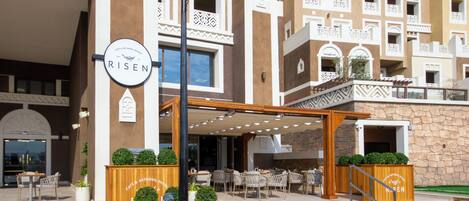 A hotel entrance with a sign reading "RISE," outdoor seating area with wicker chairs, and a stone facade.