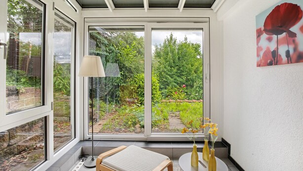 A conservatory with large windows, a chair, a table, and a vase with flowers. - Apartment 'Burgblick' with Shared Garden and Wi-Fi (Bad Urach)