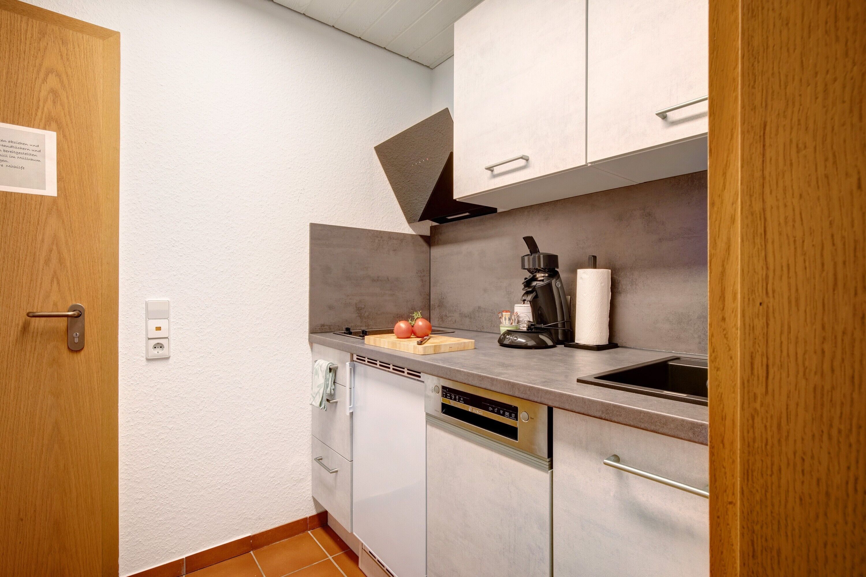 A compact kitchen with a built-in dishwasher, a coffee maker, and a cutting board with tomatoes.