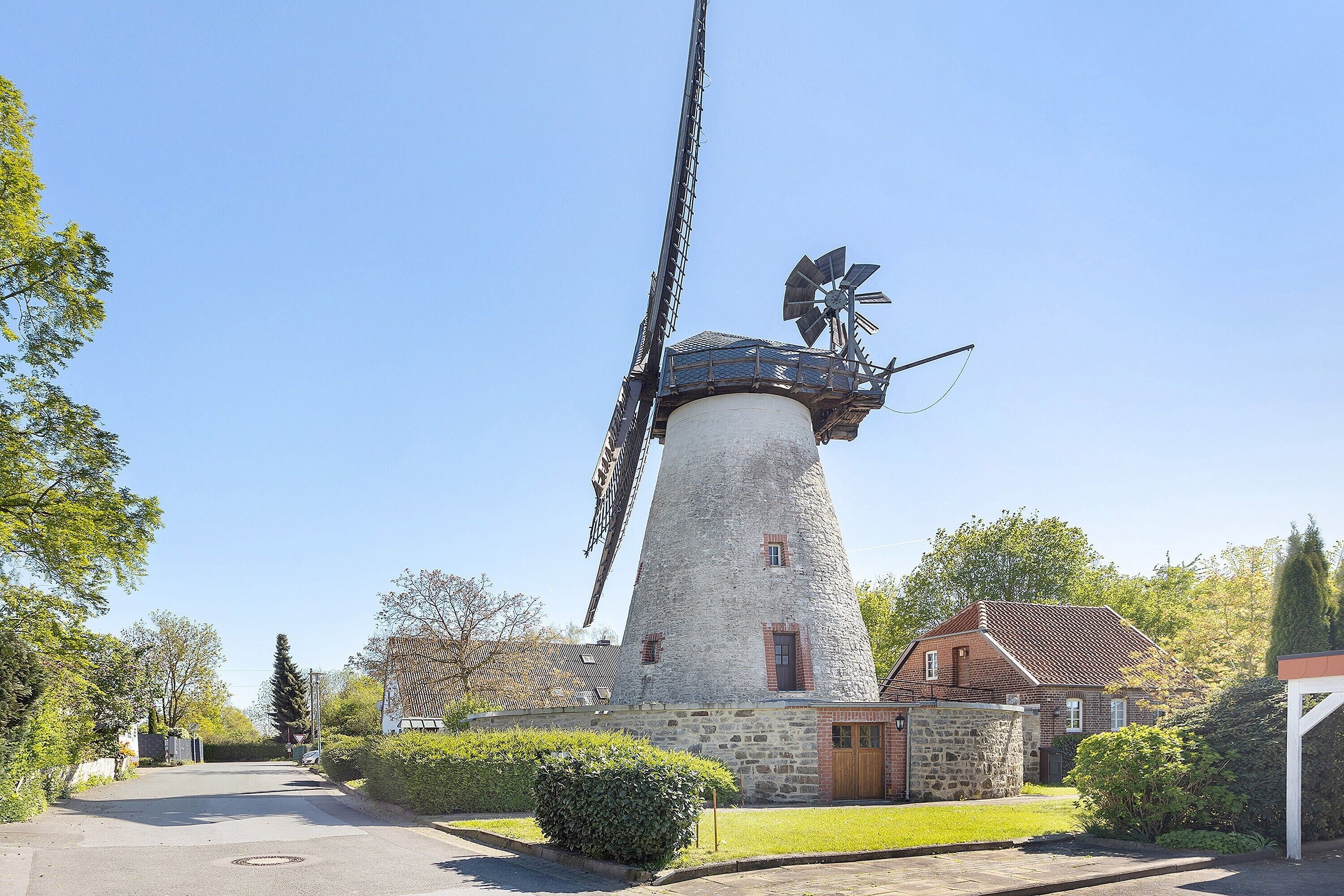 Un moulin à vent traditionnel, avec une base en pierre et un treuil métallique, entouré d’un jardin bien entretenu et d’un immeuble d’habitation.