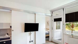 Una habitación moderna con una televisión de pantalla plana montada en una pared blanca, una cama con cabecero de madera, una ventana con persianas y una mesita con una cesta.