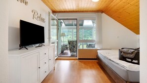 A room with a TV on a white cabinet, a bed with a blanket, a wooden ceiling, and a view through a glass door.