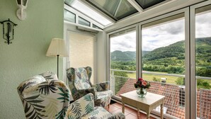 A conservatory with a view of green hills, a patterned armchair, a table with a vase, and a wall-mounted lamp.