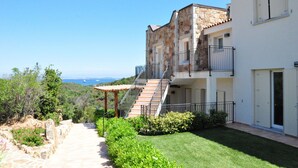 A two-story stone building with a balcony, a stone staircase leading to the upper floor, and a view of the sea in the background.