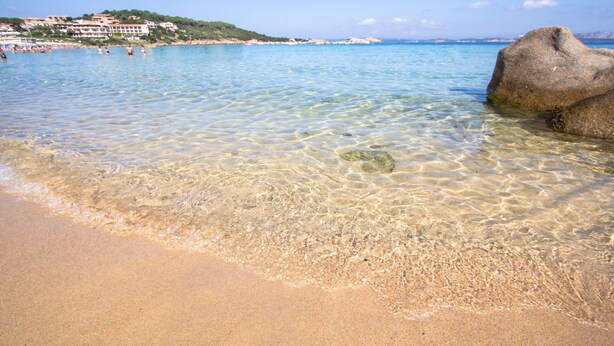 A clear beach with visible rocks and a distant building on a hill.