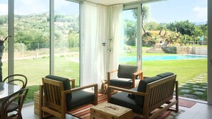 Une pièce dotée d’une grande porte-fenêtre coulissante donnant sur une piscine et un jardin. On y trouve des chaises en bois et une table avec des coussins.