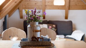 A rustic wooden interior with a metal vase of flowers on a table, wooden chairs, and a sofa in the background.
