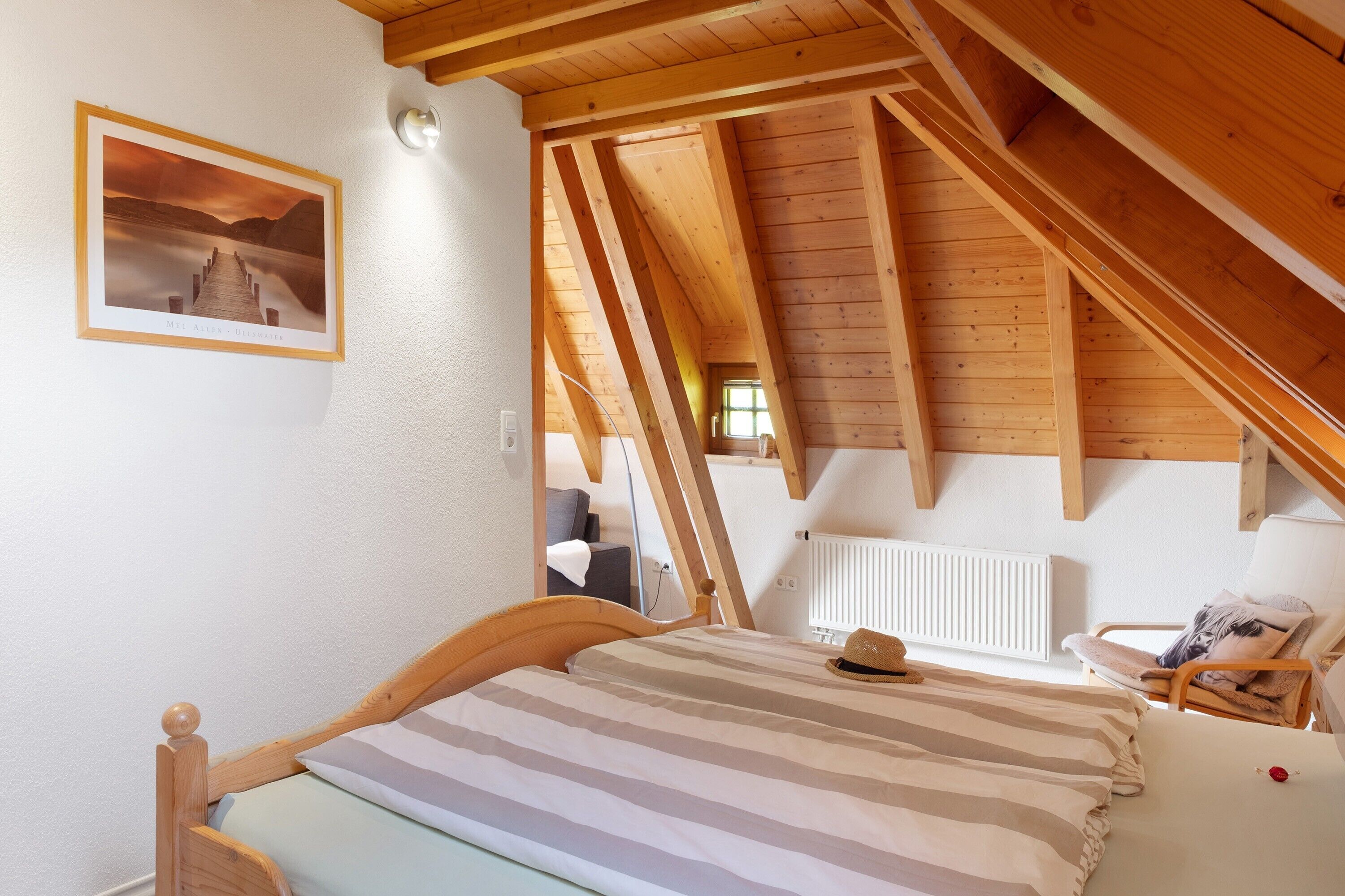 A bedroom with a wooden ceiling, a bed with striped bedding, a hat on the bed, a chair, and a radiator.