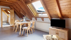 A room with a sloped ceiling, wooden beams, a skylight, a dining table with chairs, a television on a wooden cabinet, and a radiator.