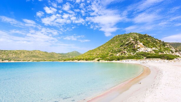 A beach with clear turquoise water, white sand, and a green hill in the background.