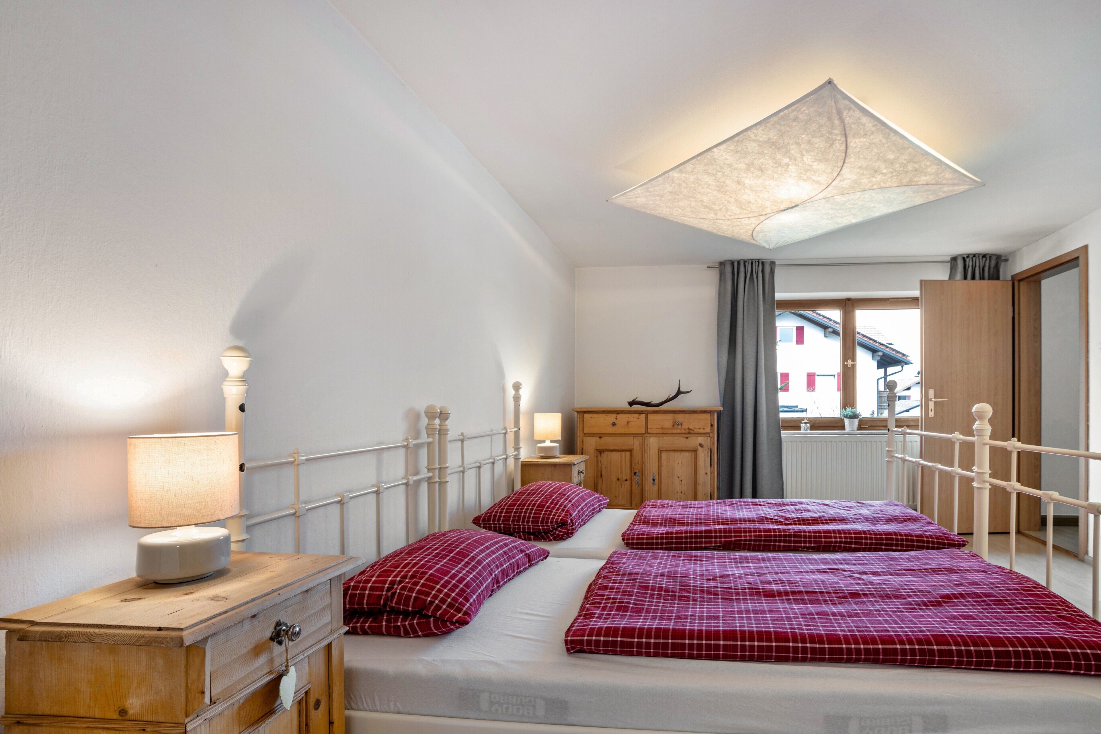 Camera da letto con due letti, una cassettiera in legno, un comodino, una lampada e vista su un edificio dalla finestra.