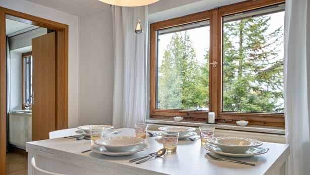 A dining area with a table set for four, a window with a view of trees, and a wooden door. - Holiday Apartment 'Silvia' with Mountain View, Shared Garden, and Wi-Fi (Rosshaupten)
