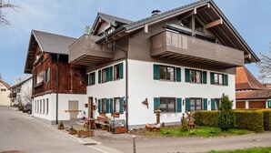 A two-story house with a sloped roof, wooden exterior, and a front porch with outdoor seating. - Holiday Apartment 'Silvia' with Mountain View, Shared Garden, and Wi-Fi (Rosshaupten)
