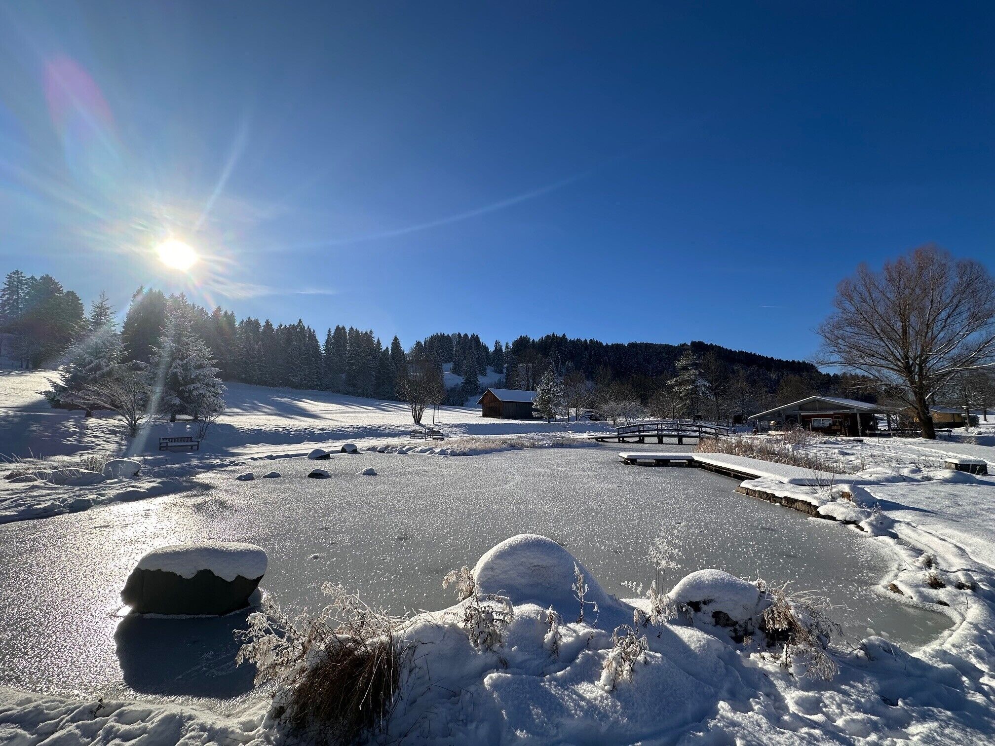 A snowy landscape with a wooden cabin, a bench, and a small bridge.