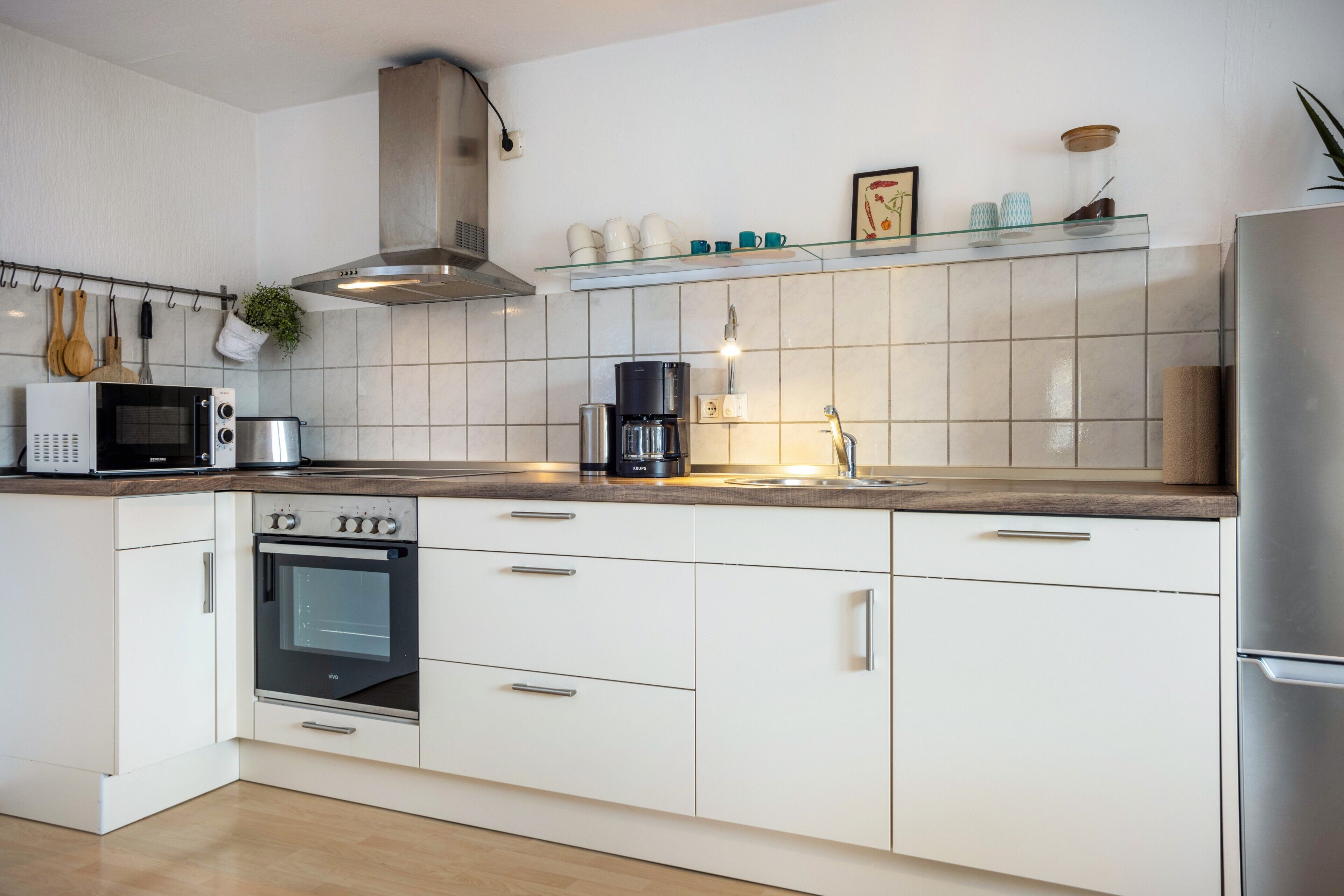 A modern kitchen with white cabinetry, a built-in oven, a microwave, a coffee maker, and a stainless steel range hood.