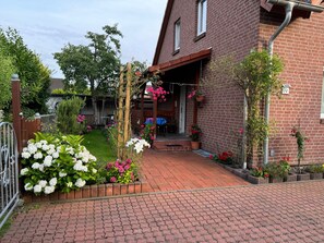 Una casa de ladrillo con entrada pavimentada, cerca de madera y jardín con diversas plantas y flores.