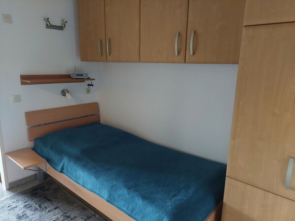 A single bed with a teal mattress, wooden headboard, and a small bedside table. There is a wooden wardrobe above the bed with two doors and a shelf.