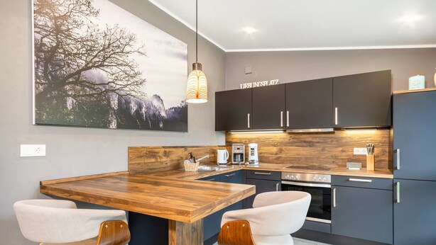 Une cuisine moderne avec un îlot central en bois, deux tabourets de bar, des meubles de rangement sombres et un grand tableau accroché au mur.