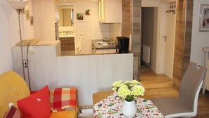 Een compacte leefruimte met een kitchenette, een eethoek met een tafel en bloemen, en een bank met een rode kussens.