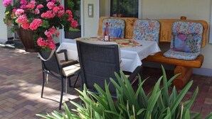 Een overdekt terras met een tafel voor twee, een bank met bloemenkussens en een hangende plantenbak met roze bloemen.