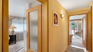 Un corridoio con una porta in legno, una lampada, un quadro incorniciato e vista su un'altra stanza con una sedia.