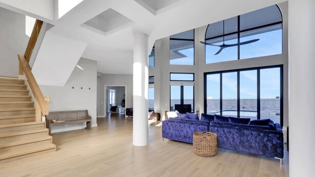 A spacious living area with a wooden staircase, a large sofa, and a view of the sea through large windows. - Luxury River View Estate | Heated Pool & Amazing Sunsets | Perfect Gathering for Families & Friends (Fort Myers)