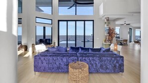 A spacious living room with a large sectional sofa, a ceiling fan, and a view of the sea through large windows. - Luxury River View Estate | Heated Pool & Amazing Sunsets | Perfect Gathering for Families & Friends (Fort Myers)