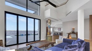 A spacious living area with a large window overlooking a body of water, a modern kitchen in the background, and a cozy seating area with a unique wooden coffee table. - Luxury River View Estate | Heated Pool & Amazing Sunsets | Perfect Gathering for Families & Friends (Fort Myers)