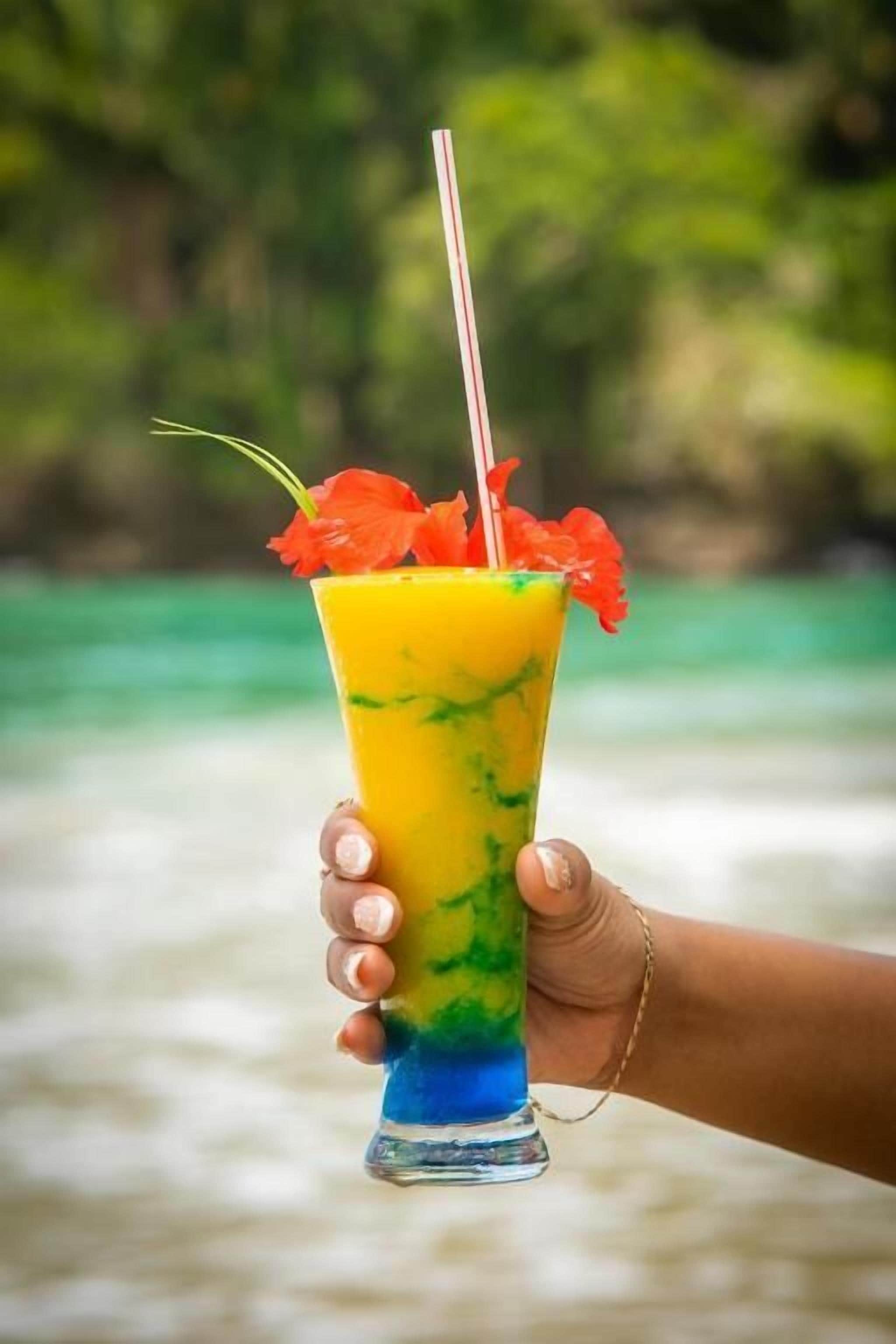 A hand holding a tall glass with a layered drink, a straw, and a flower garnish, against a blurred natural background.