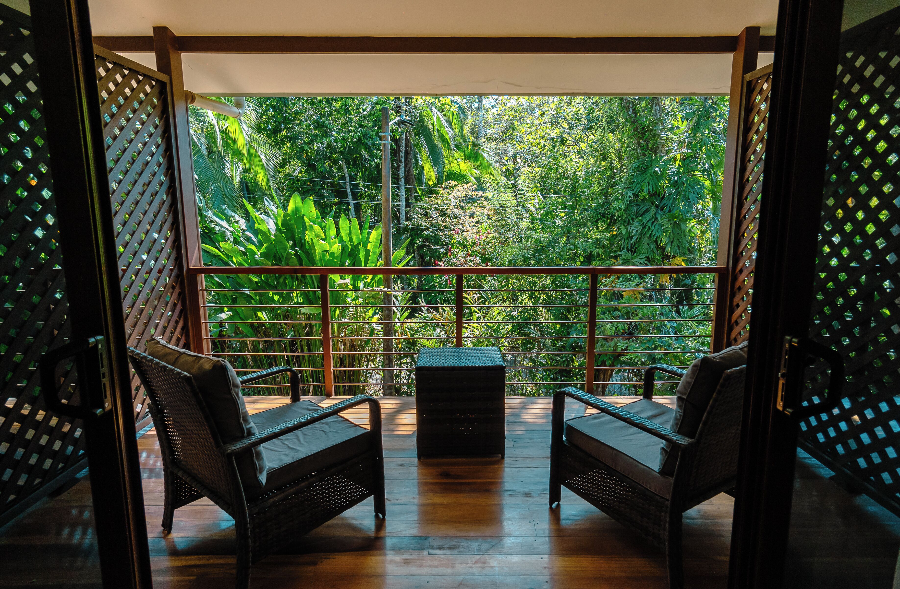 A balcony with two wicker chairs and a small table, surrounded by a wooden floor and a metal railing, overlooking a lush green garden.