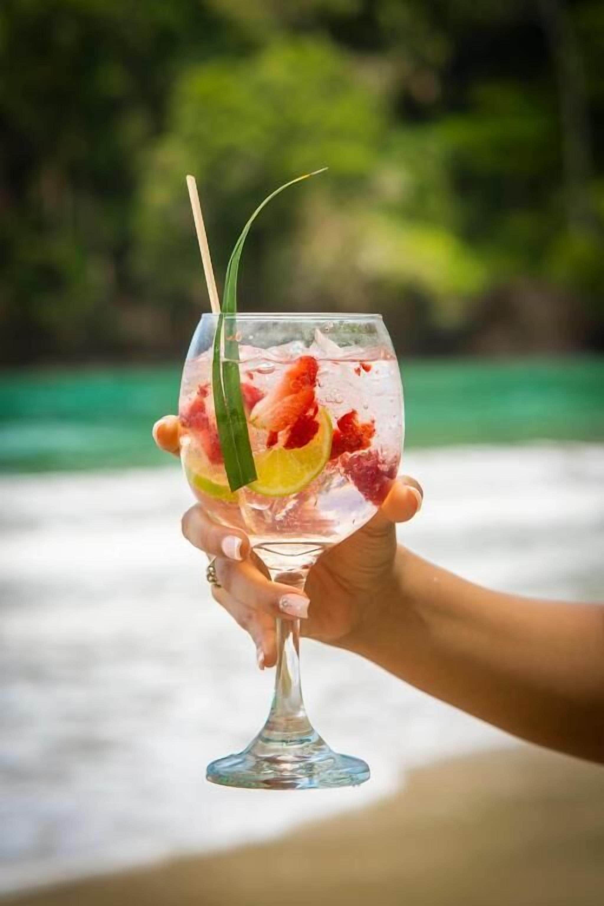 A hand holding a clear glass with a drink garnished with fruit and a straw.