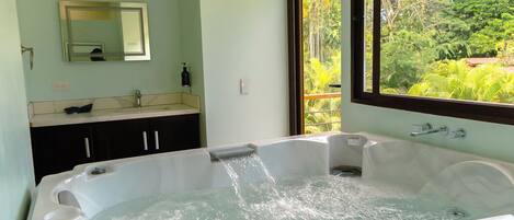Un baño con una bañera grande, un lavamanos, un espejo y una ventana con vistas al follaje.