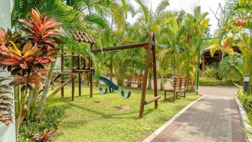 A playground with swings and a slide, surrounded by palm trees and a brick pathway.