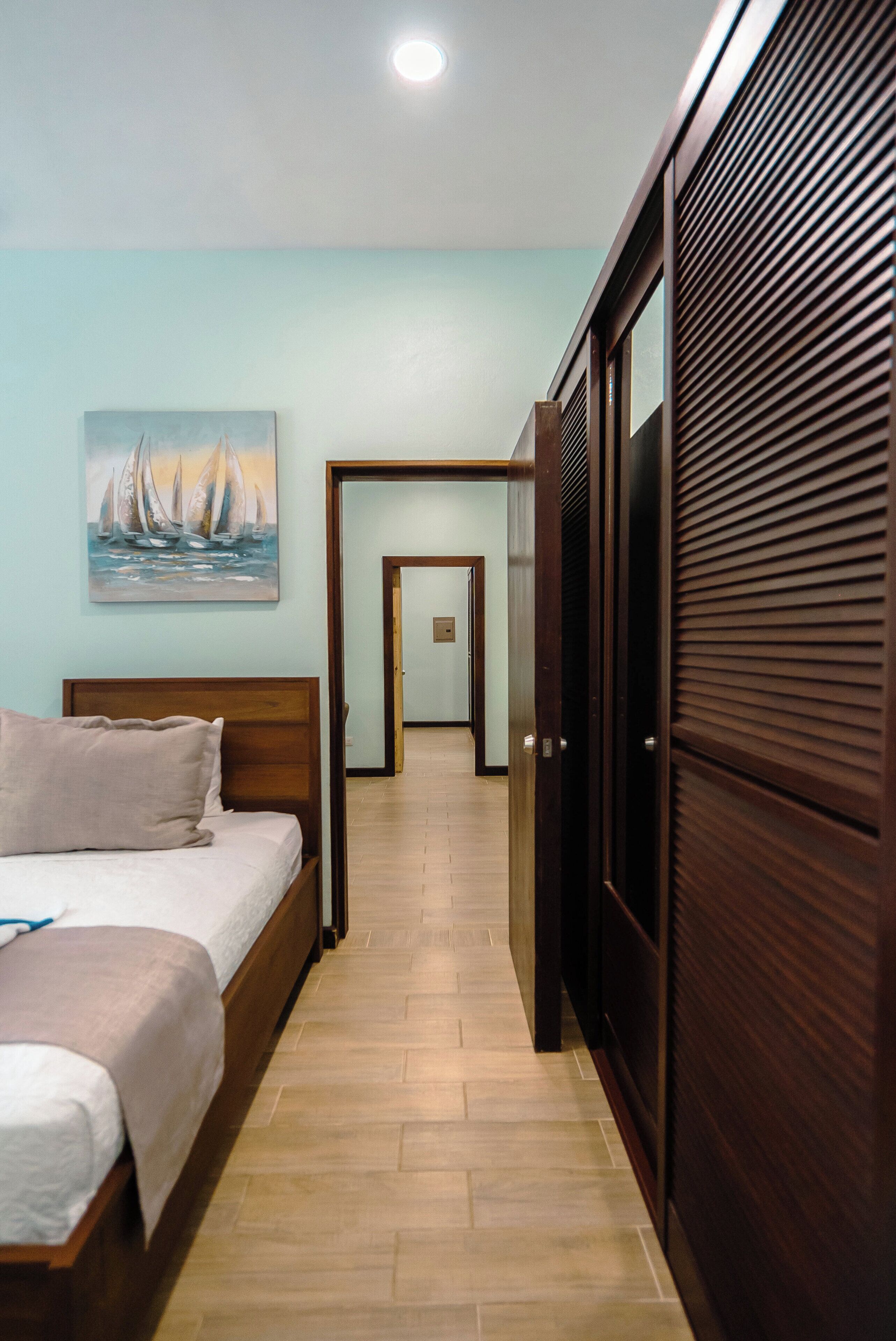 A hotel room with a bed, a painting on the wall, a wooden wardrobe, and a hallway leading to another room.