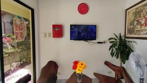 Ein Zimmer mit einer Uhr, einem Fernseher, einer roten Notfallbox, einem Ventilator und einer Vase mit Blumen.