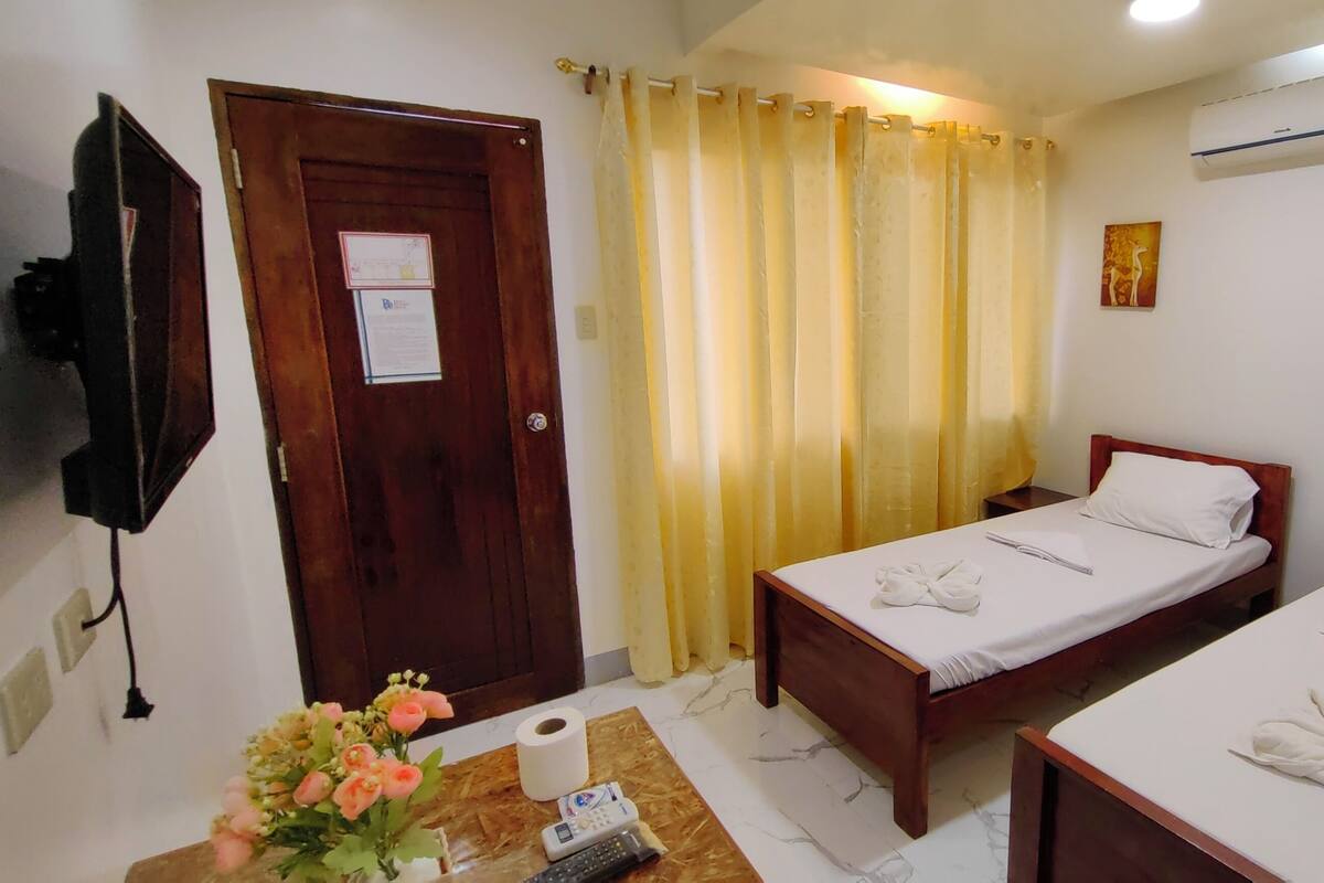 A hotel room with two beds, a wooden door, a curtain, a wall-mounted TV, a painting, and a small table with a vase of flowers.