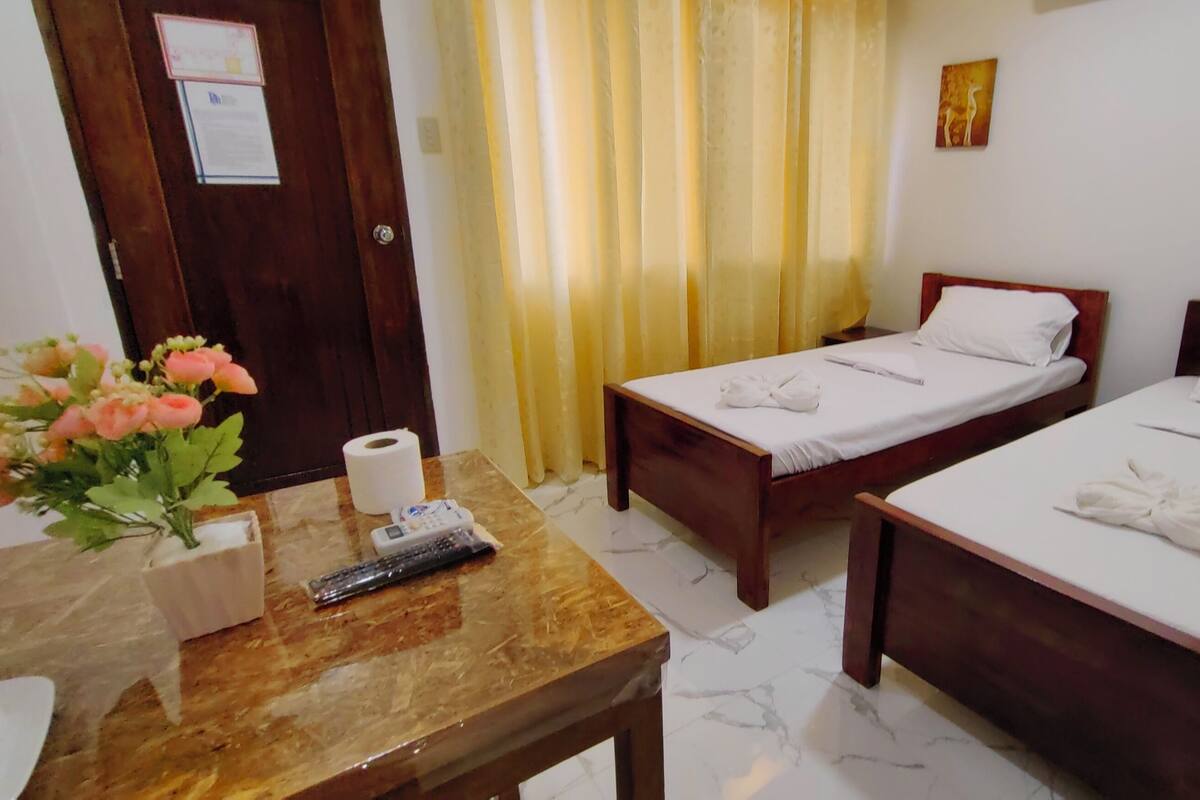 A hotel room with two single beds, a wooden table with a vase of flowers, a remote control, and a toilet paper roll.